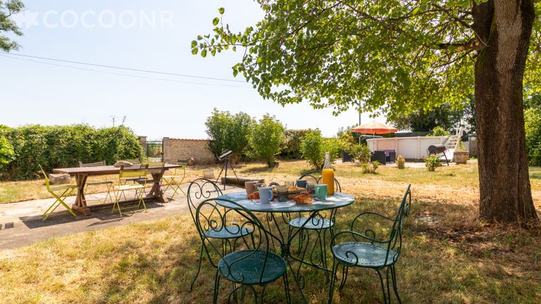 Le jardin de La Maison du Tonnelier : une table en fer forgé dressée à l'ombre d'un grand arbre, une seconde table en bois sur la terrasse, et la piscine hors-sol au fond du terrain.